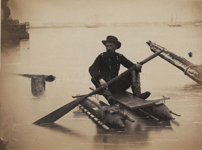 A man in historical clothing paddles a raft, demonstrating an invention ahead of its time on a calm body of water.