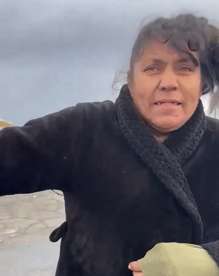 A person outdoors in a black coat, reacting to something off-camera, under a cloudy sky.