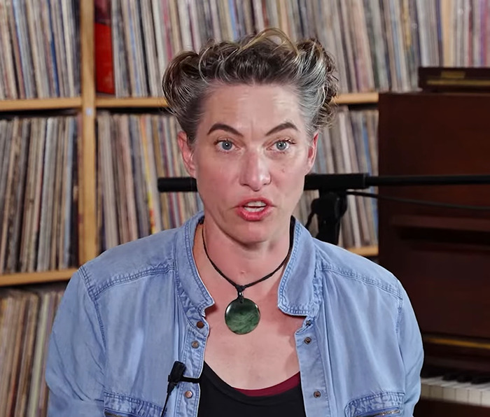 Person sitting in front of shelves filled with records, speaking, and wearing a blue denim shirt and necklace.