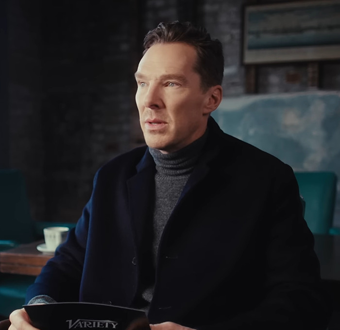 Benedict Cumberbatch seated indoors wearing a dark coat and gray turtleneck.