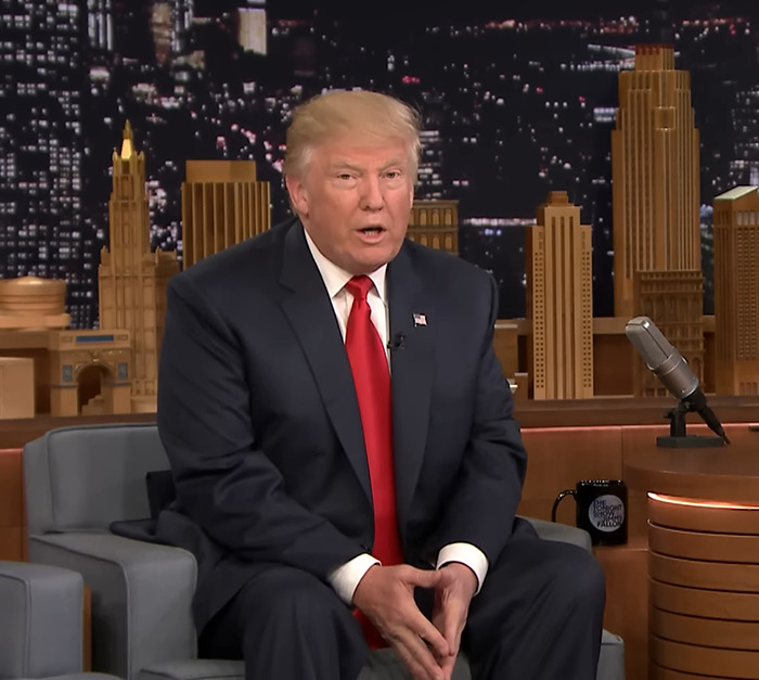 Donald Trump in a dark suit sitting and speaking in front of a cityscape backdrop on a talk show set.