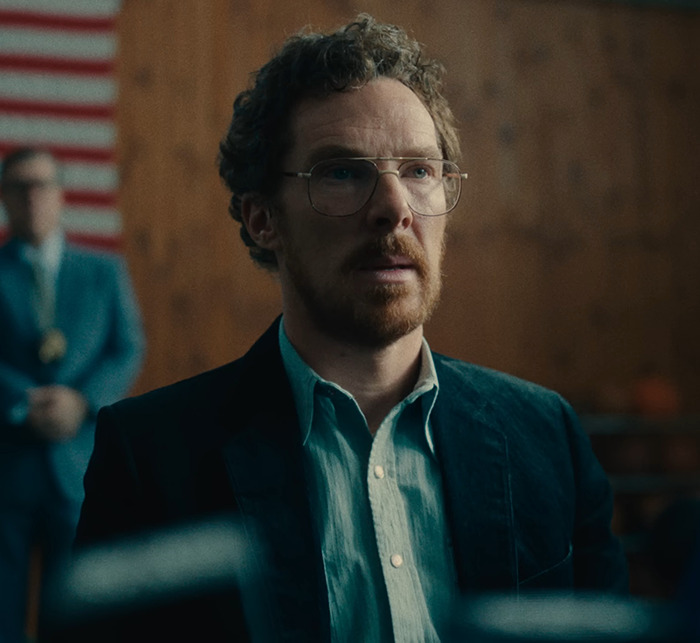 Benedict Cumberbatch in a suit, sitting indoors, with an American flag in the background on a scene from 'Eric.