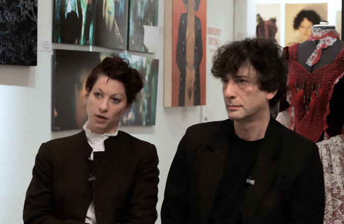 Two people sitting in an art gallery, dressed in dark clothing, surrounded by paintings and a mannequin with a red garment.