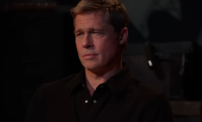 A man in a black shirt appears solemn, with a blurred dark background.