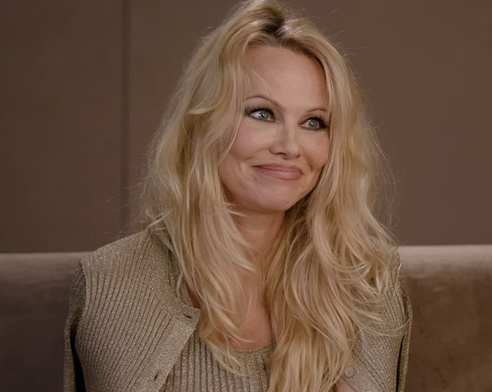 Blonde woman smiling, wearing a shimmery top, against a neutral background.