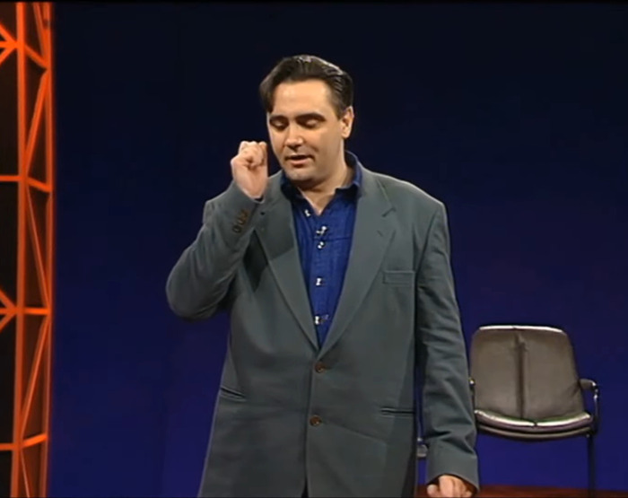 Tony Slattery performing on stage in a suit with a chair in the background.