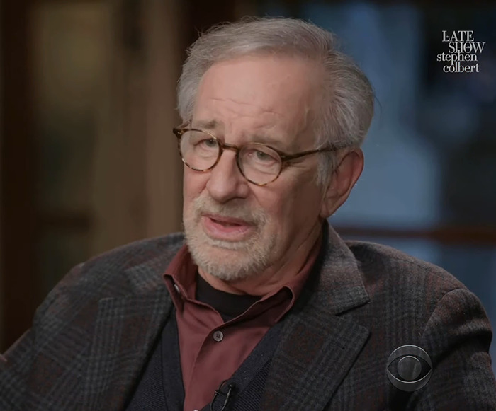 An older man with glasses discusses tributes from Hollywood, wearing a plaid jacket and maroon shirt on a talk show set.