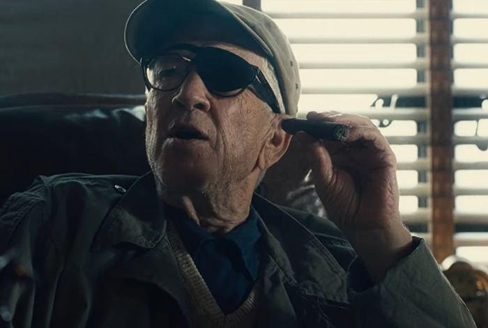 A man with an eye patch and cap, holding a cigar, sits in a dimly lit room with blinds in the background.