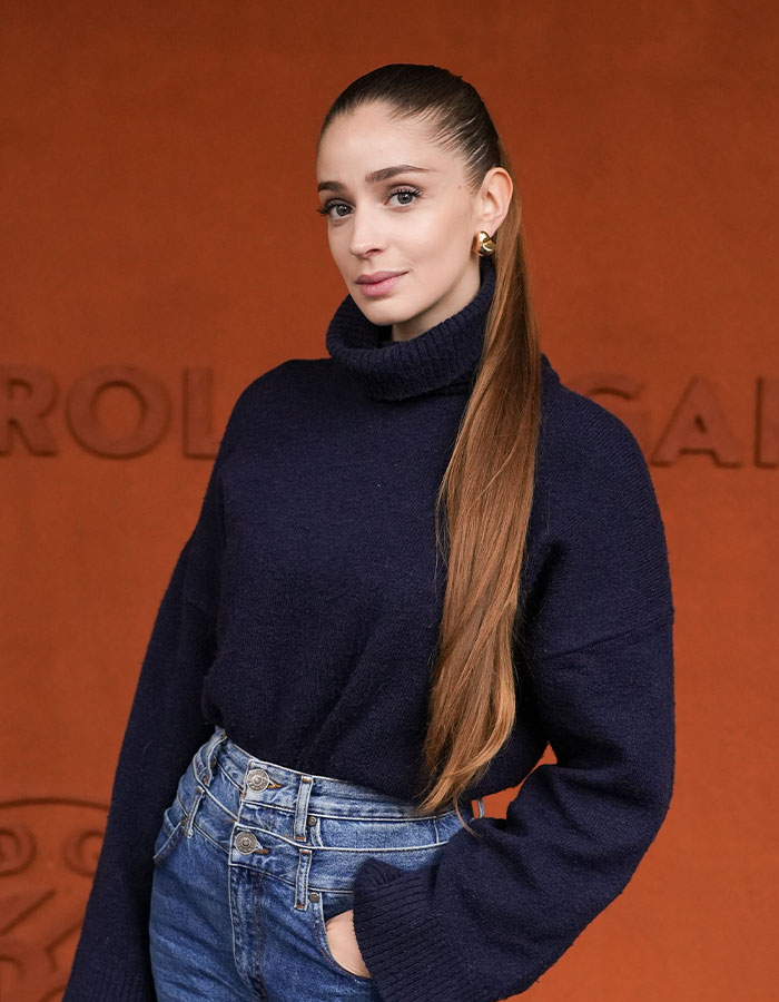 Ballerina posing against orange background, wearing a navy sweater and jeans.