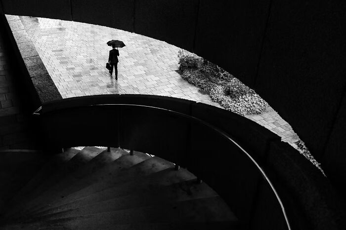 Black and white photo of a person with an umbrella on a walkway, viewed from a spiral staircase. Artist Gallery Awards.