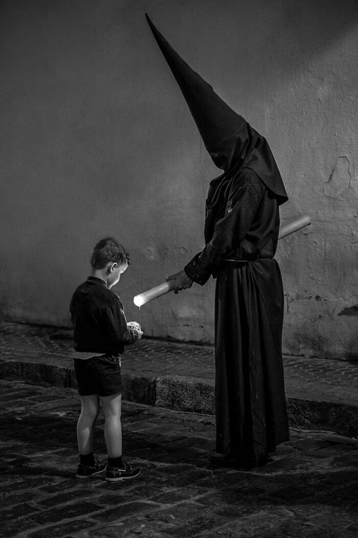 A child stands opposite a hooded figure holding a candle in an award-winning black-and-white photo from The Artist Gallery Awards.