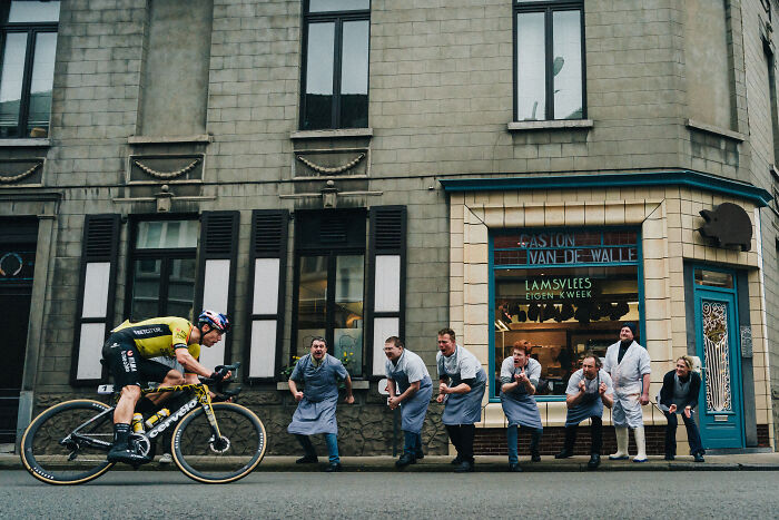 Cyclist competing on a city street as a group of cheering onlookers in aprons encourage him, showcasing sports photography.