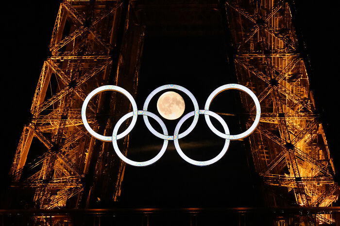 Olympic rings with full moon framed within, illuminated against the Eiffel Tower, capturing world sports photography magic.