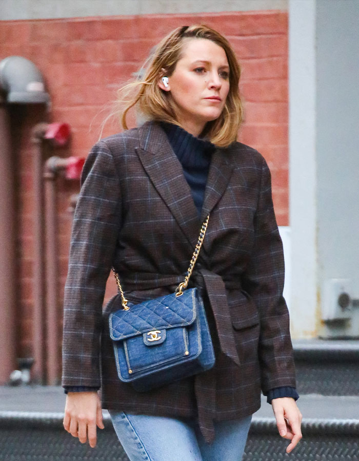 Person in a plaid jacket, wearing earbuds and a denim shoulder bag, walking near a brick wall.