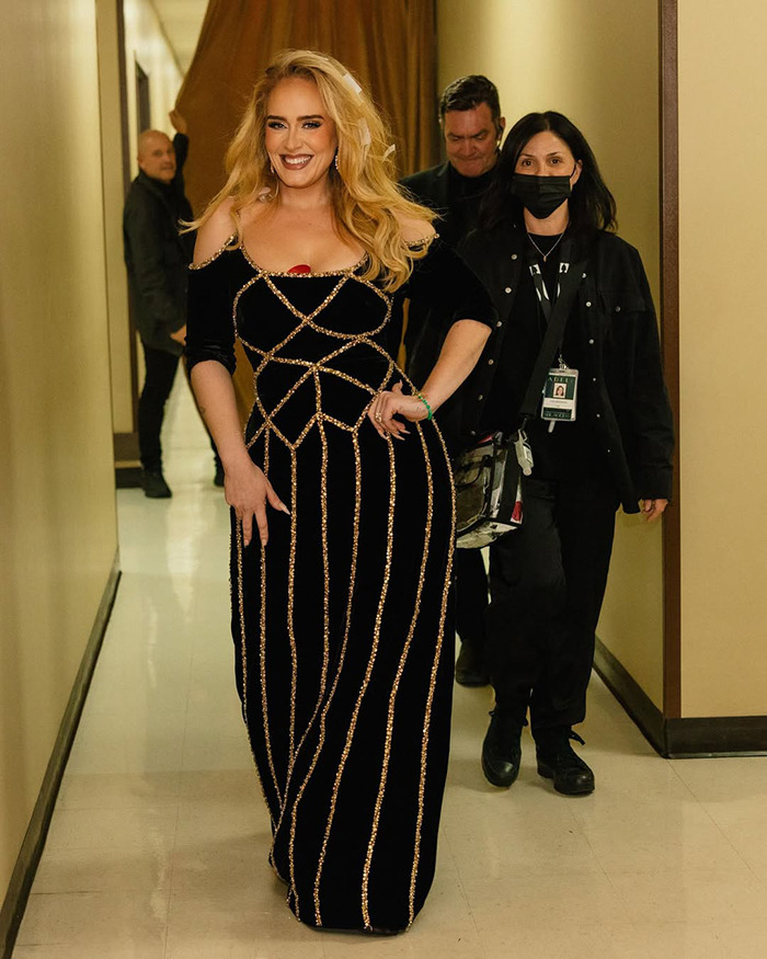 Woman in an elegant dress, smiling and walking in a hallway with others, sparking pregnancy speculation.