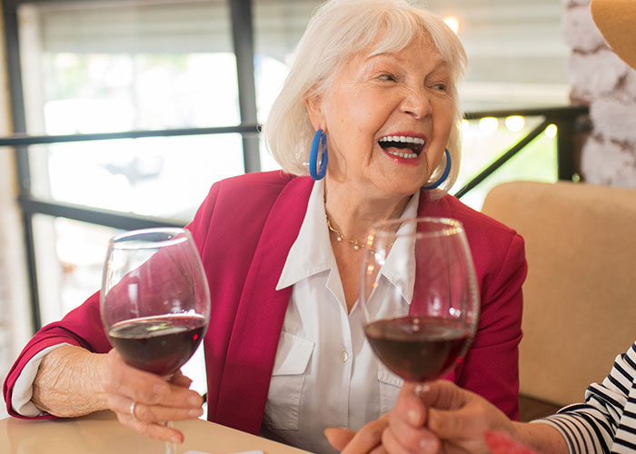 Elderly woman in a pink blazer laughing and holding a glass of red wine at a social gathering.