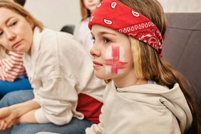 Child wearing a red bandana with a flag painted on cheek, sitting next to a woman, illustrating kids feeling like foreigners.