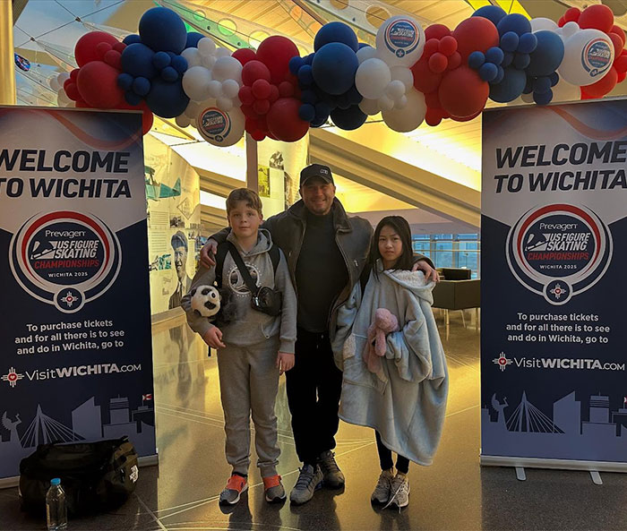 Young ice skating duo with an adult at Wichita event entrance, surrounded by colorful balloons.