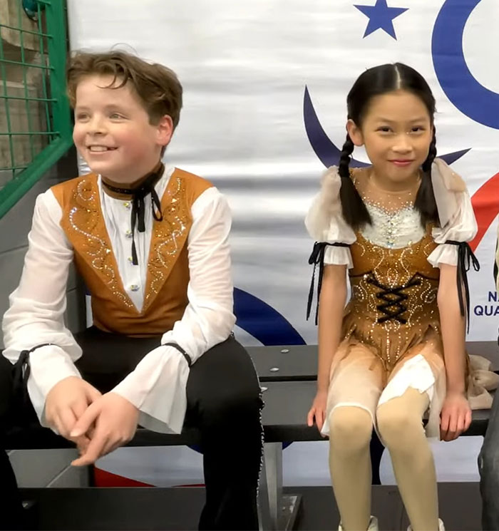 Adorable child ice skating duo sitting together in matching costumes on a bench.