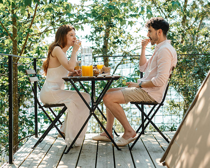 A couple dining outdoors, enjoying a breakfast with juice, surrounded by lush trees on a wooden deck.