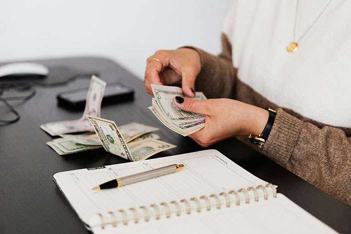 Counting money at a desk, a person plans finances for a bachelorette mini-vacation, with a notebook and pen nearby.