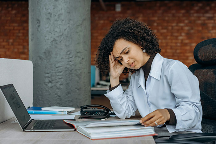 A woman looks stressed while working at a desk, possibly dealing with bachelorette mini vacation drama, near a laptop and documents.