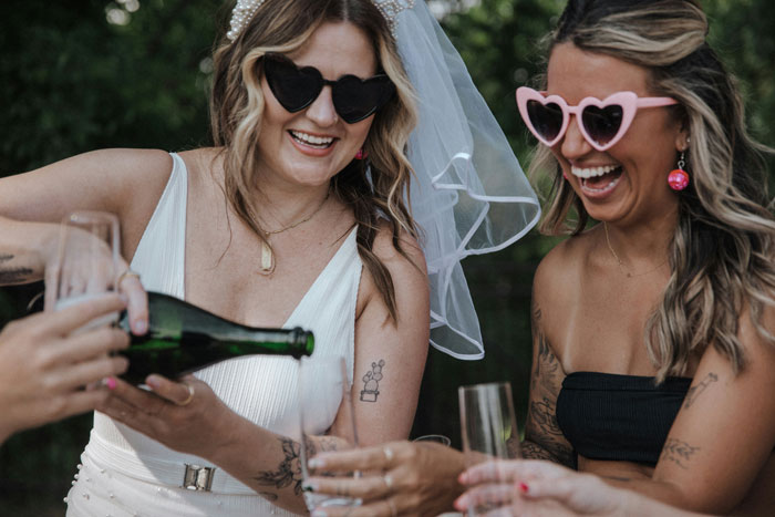 Brides at a bachelorette party laughing and pouring champagne, wearing heart-shaped sunglasses.