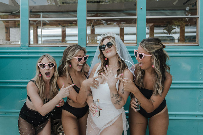 Women in swimsuits with heart-shaped sunglasses celebrate at a bachelorette party, posing playfully in front of a blue bus.