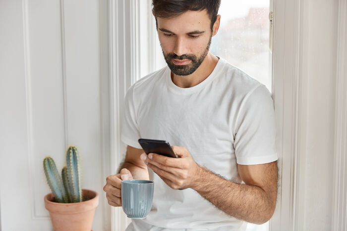 Man in white shirt holding a mug, looking at his phone, sitting by a window next to a cactus.