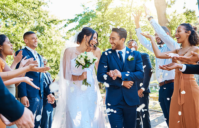 Bride and groom smiling under confetti, surrounded by guests celebrating their wedding day outdoors.