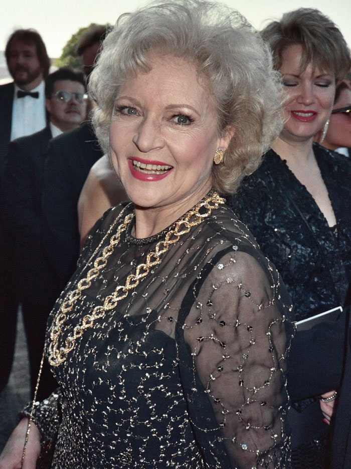 A woman in a glamorous black dress, smiling at an event, surrounded by people.