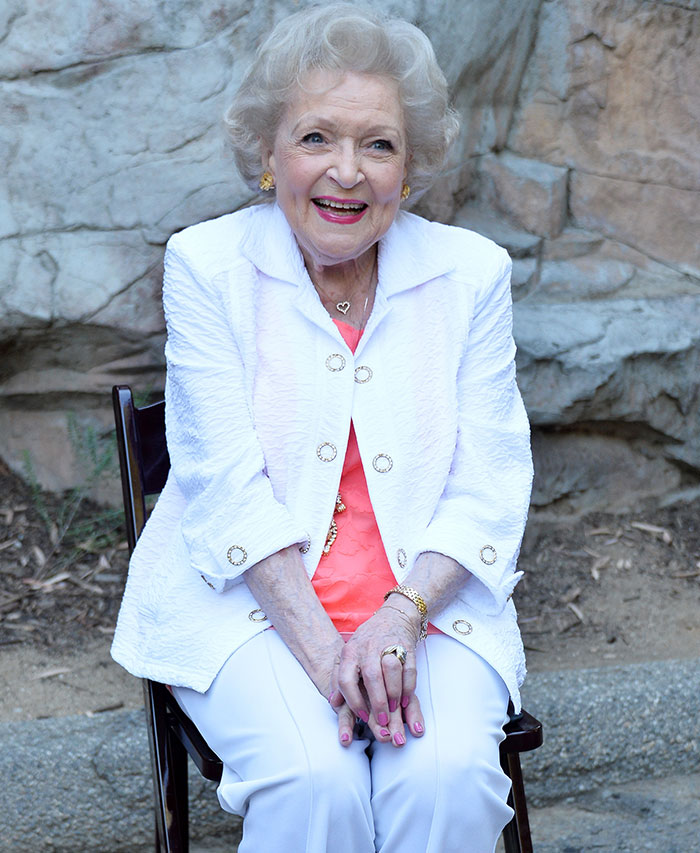 Elderly woman in a white outfit sitting outdoors, smiling warmly at the camera.