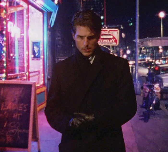 A celebrity walking at night in a city, wearing a dark coat and gloves, with neon lights and traffic in the background.