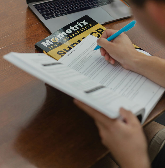 "Person studying, taking notes with a pen, referencing Mometrix test prep book, focusing on common scams in America."