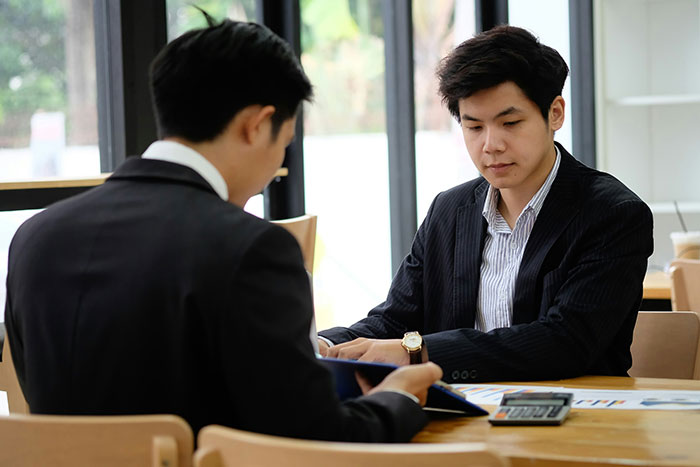 Two men in suits discussing documents, potentially involving common scams in America, seated indoors at a table with a calculator.