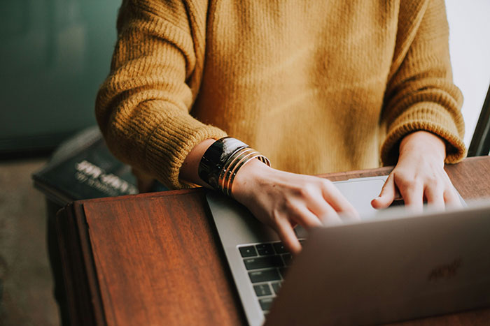 Person typing on a laptop, researching common scams in America, wearing a mustard sweater and bracelets.