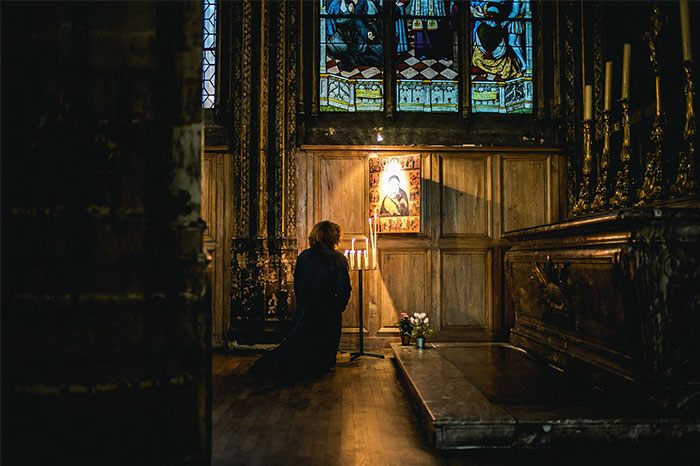 Person kneeling in a dimly lit church, reflecting on history, in front of a lit candle and religious icon.