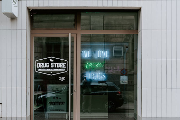 Glass storefront with "The Drug Store" logo and a neon sign saying "We love legal drugs," reflecting car and street.