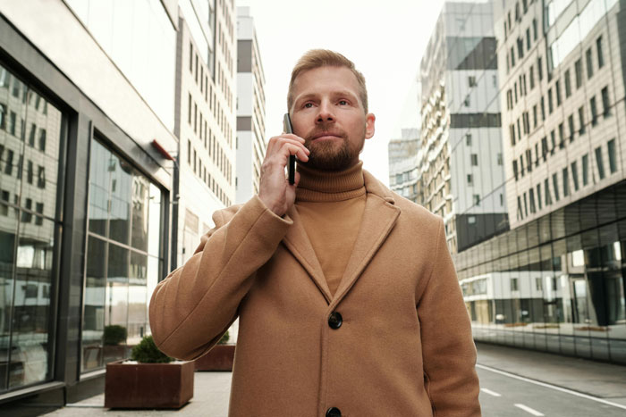 Man in tan coat talking on phone while standing on a city street, focused on an emotional conversation.