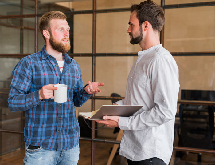 Two men in conversation, one holding a laptop and the other a mug, discussing wedding dress shopping.