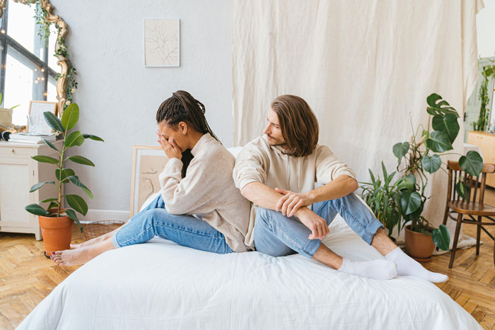 A couple sitting back-to-back on a bed, looking upset during wedding dress shopping day.