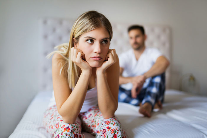 Woman looking upset on a bed, with a man sitting in the background, wedding dress shopping keywords.