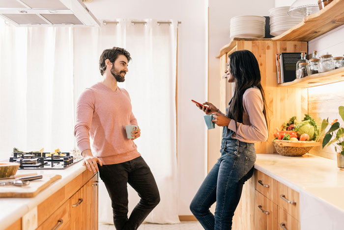 Man and sister-in-law talking in a cozy kitchen, both holding mugs, with fruit and spices on the counter.