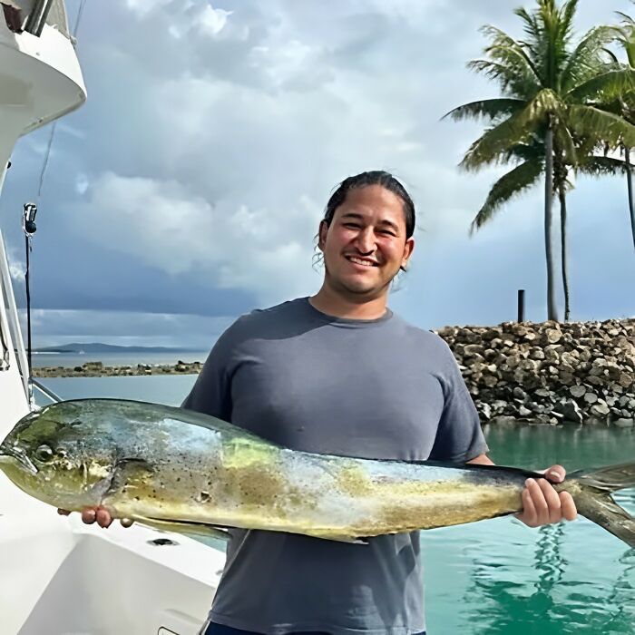 Man smiling while holding a large fish on a boat, with palm trees and rocky shoreline in the background.