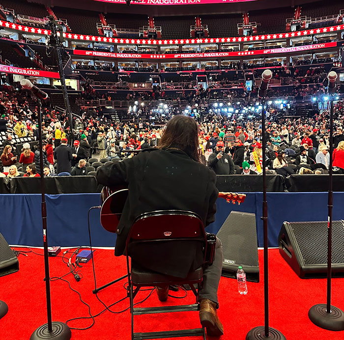 Musician at inauguration, viewed from behind on stage, with crowd and bright lights in auditorium.