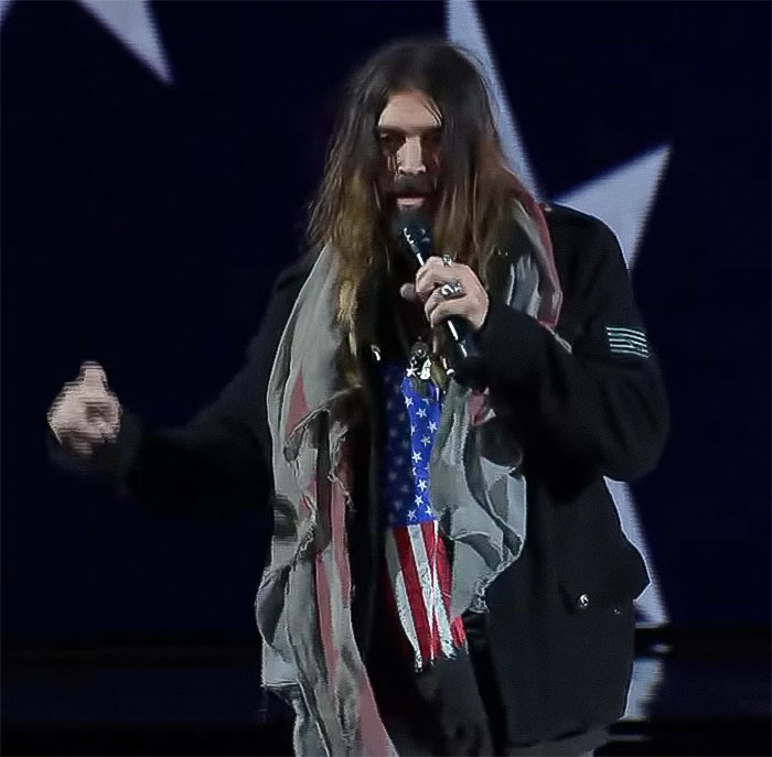 Man holding a microphone, wearing a star-patterned scarf with long hair during a performance.