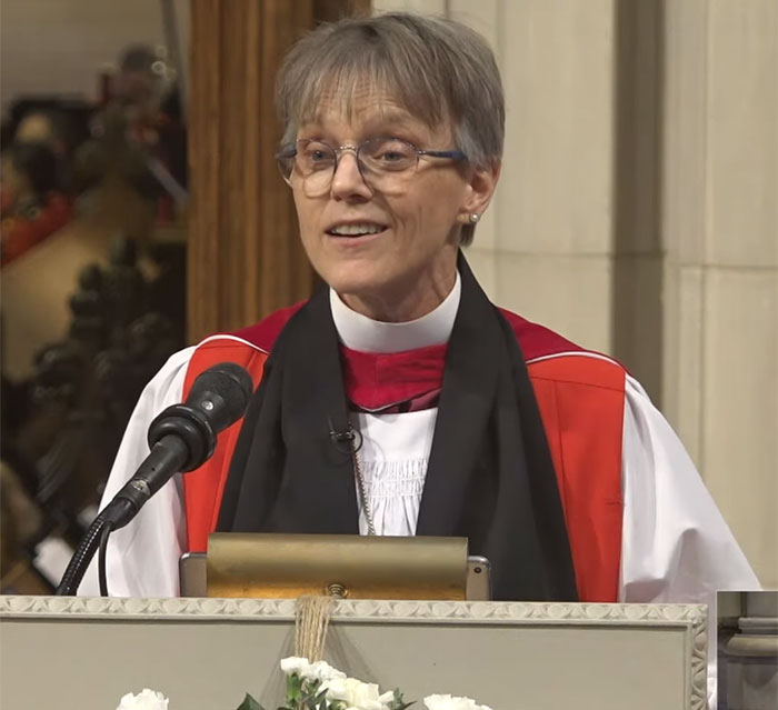 Bishop addressing audience, wearing clerical attire, at a podium with microphone in a church setting.