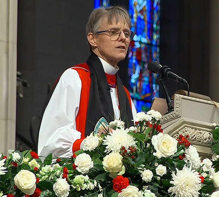 Bishop delivering a sermon at a podium adorned with flowers, addressing Americans.