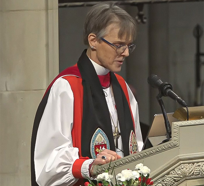 Bishop in ceremonial robes delivering a sermon at a lectern with a microphone, emphasizing a message to Americans.