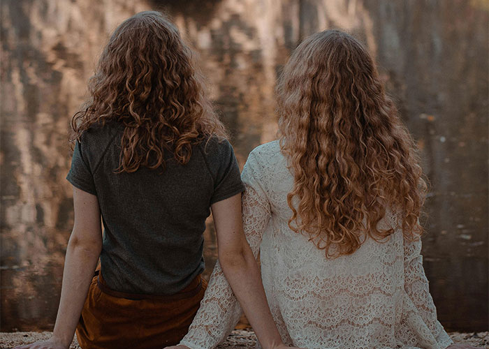 Two people with curly hair sitting by a reflective water surface, possibly discussing unsolved mysteries.